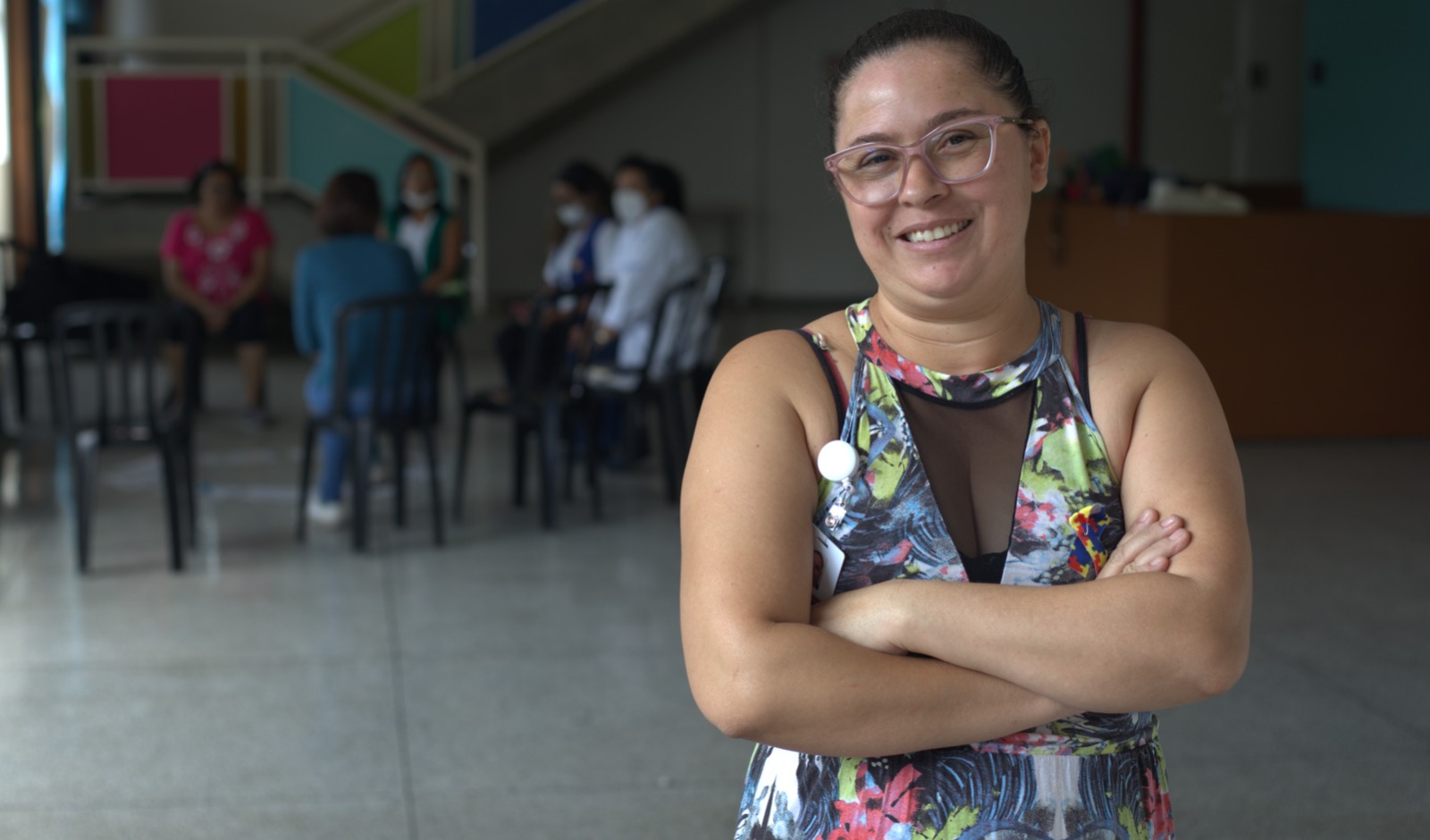 Na imagem, Deusa está sorrindo e com os braços cruzados. Ela usa uma regata estampada, óculos e está com o cabelo preso. Ao fundo, há mais cinco pessoas sentadas em cadeiras em círculo, com os rostos desfocados